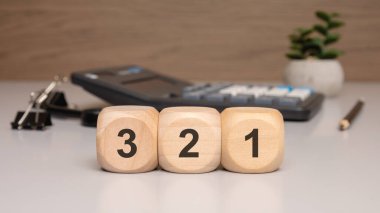 3, 2, 1 numaralı tahta bloklar, geri sayım sırasına göre dizilmiş, arka planda hesap makinesi olan modern bir ofis masasına yerleştirilmiş.