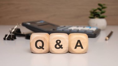 Tahta bloklar Q A 'yı hesap makinesi, kalem ve küçük bitkiyle temiz bir ofis masasına yerleştirerek iş ortamı veya finans ortamında soru ve cevap kavramını sembolize ediyor.