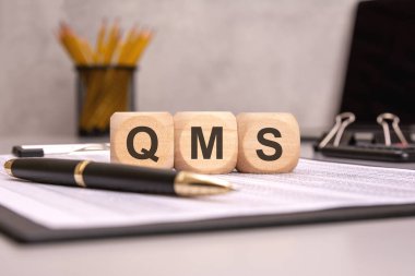 Ofis çalışma alanındaki kağıt üzerinde QMS kısaltması olan tahta bloklar. Kalite yönetim sistemi, ISO standartları ve iş süreci iyileştirmesinin sembolü.