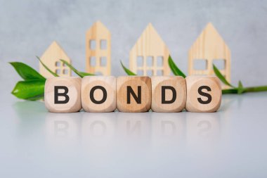 Bloklar, dekoratif yapraklar ve ahşap ev siluetleriyle ışık masasında Kelime BONDS oluşturur. Yatırım menkul kıymetlerini, borsa finans ekonomisini ve iş stratejisini sembolize eder