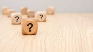a natural wooden block printed with question mark sits sharply in the foreground on a wooden desk, while other cubes blur in the background. symbolizes uncertainty, inquiry, decision-making, questions.