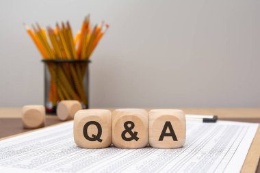 wooden blocks reading Q A placed on a printed table in focus, with pencils and blurred background. Suitable for Q A, interviews, FAQs, support, discussion.