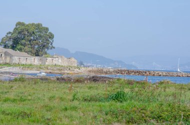 Sahildeki tarihi bir taş binanın manzarası. Etrafı dağlarla çevrili yeşil bir bina ve uzakta mavi bir gökyüzü. Huzurlu deniz manzarası seyahat temaları için ideal.