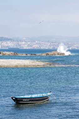 Küçük mavi balıkçı teknesi sakin sahil suyuna demirlemiş. Uzak şehir silüeti ve kayalıklardaki dalgalar seyahat ve turizm kavramları için huzurlu yaz atmosferlerini çağrıştırıyor..