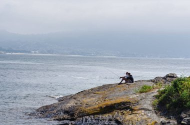 Kayalık bir kıyı şeridinde balık tutan bir adam, deniz kenarında sakin bir anın tadını çıkarırken, arkasında dağlar ve doğa vardı. Rahatlama, rahatlama ya da açık hava yaşam tarzı kavramları için ideal..