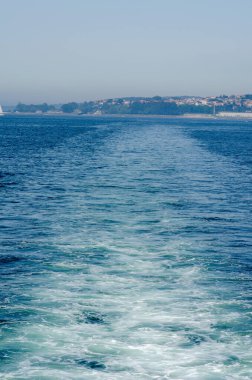 Sakin mavi okyanus suları boyunca uzanan köpüklü bir gemi izi berrak bir gökyüzü altında manzaralı bir sahil kasabasına doğru uzanıyor. Seyahat ve sükuneti çağrıştırıyor..