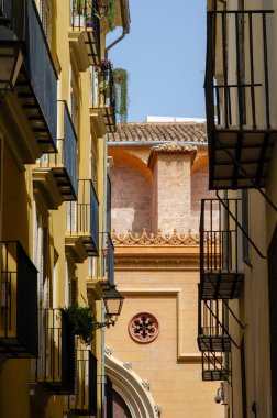 Süslü taş işçilikli, güneşli tarihi bir binanın resmi. Klasik sarı cephelerle çevrili dar bir dar sokaktan görülüyor ve İspanya 'da demir balkonlar inşa ediliyor..