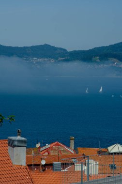 Sakin mavi bir deniz manzaralı turuncu çatılar. Yelkenliler ve uzak yeşil tepeler. Huzur ve yaz rahatlığını çağrıştırıyor..