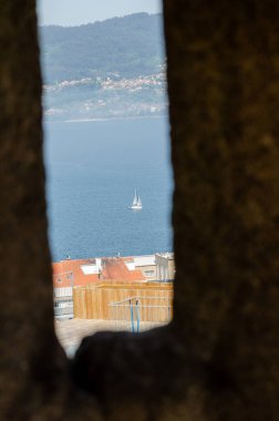Derin mavi denizde yüzen barışçıl yelkenli, tarihi bir taş pencereden köyün çatıları ve uzak ormanlık tepeleri ile görülüyor. Sahil gezisi manzarası.