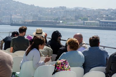 Çeşitli bir grup insan, açık bir güvertede, deniz manzaralı bir nehir gezisi sırasında dinleniyor ve sohbet ediyor, deniz kenarında canlı bir şehir manzarası var, boş zamanları ve seyahat ortamını çağrıştırıyor..