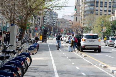 Modern bisiklet şeridi, park halindeki bisikletleri, araçları ve yayaları olan kalabalık bir Avrupa caddesi. Güneşli bir günde kentsel hareketlilik ve çevre dostu ulaşım kavramı.