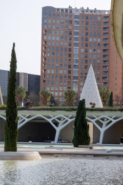 Modern yüksek katlı bir apartman bloğu, eşsiz geometrik yapılar ve halka açık bir şehir parkında yansıyan bir su havuzu içeren çağdaş şehir manzarası, sakin bir atmosferi çağrıştırıyor..