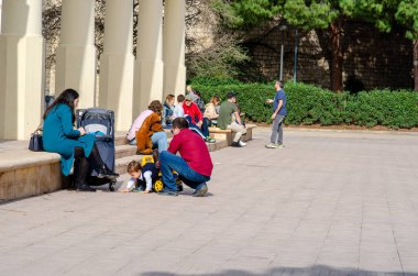Aileler ve yetişkinler şehir parkında güneşli bir günün tadını çıkarıyorlar, banklarda oturuyorlar ve rahat ve dost canlısı bir ortamda dışarıda etkileşime geçiyorlar..