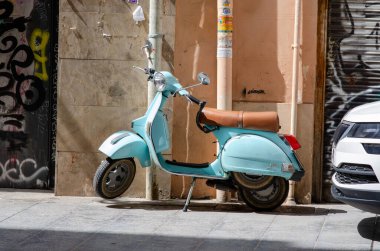 Deri koltuklu klasik retro scooter, parlak güneş ışığında bir şehir caddesinde duruyor. Akdeniz atmosferini ve şehir ulaşımını çağrıştırıyor..