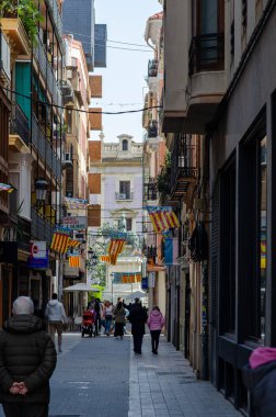 Valencia, İspanya 'da güneşli bir günün tadını çıkaran renkli Valencia bayrakları ve yayalarla süslenmiş canlı dar bir caddenin kent manzarası. Seyahat ve kültür temaları için mükemmel.