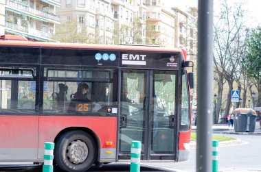 Valencia, İspanya 'da güneşli bir cadde boyunca ilerleyen kırmızı kentsel EMT şehir otobüsü içinde insanlar ve arka planda modern konut binaları.