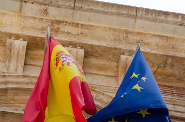 İspanya ulusal bayrağı ve AB bayrağı, Avrupa 'daki siyasi birlik ve ortaklığı simgeleyen eski bir taş binanın önünde birlikte sallanıyor.