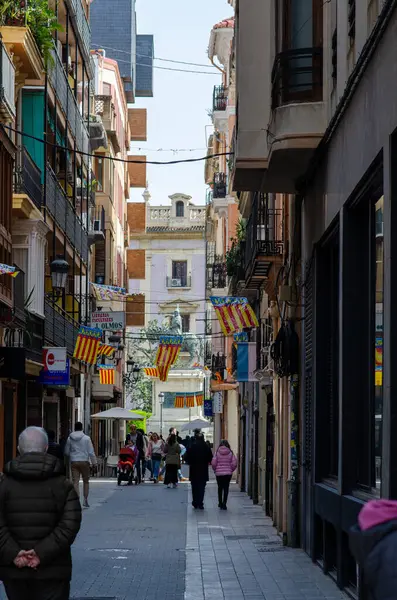 Valencia, İspanya 'da güneşli bir günün tadını çıkaran renkli Valencia bayrakları ve yayalarla süslenmiş canlı dar bir caddenin kent manzarası. Seyahat ve kültür temaları için mükemmel.