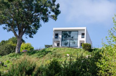 Panoramik pencereli çağdaş villa manzaralı bir tepenin üzerinde, modern mimariyi yemyeşil doğayla harmanlıyor. Gayrimenkul, seyahat ve mimari temalar için ideal.