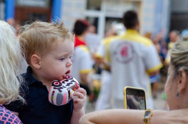 Küçük bir çocuk oyuncak bir araba tutuyor ve açık hava festivalini izliyor, bir kadın akıllı telefonuyla anı yakalıyor. Aile gezisi, gizli şehir hayatı, çocukluk merakı..