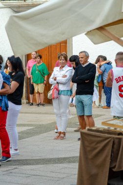 Farklı yaşlardan bir grup insan açık hava pazarı veya festivalinde sırada bekliyor. Sahne, şehir yaşamını ve sosyal etkileşimi rahat bir atmosferde yakalıyor..