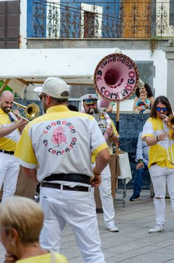 Canlı sokak bandosu bir açık hava festivalinde kalabalığı eğlendiriyor. Sarı ve beyaz kıyafetli müzisyenler enstrüman çalıyor, festival enerjisi ve topluluk ruhunu yakalıyorlar..