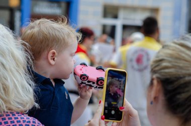Genç bir çocuk, canlı bir şehir sokak etkinliği sırasında bir yetişkin tarafından fotoğraflanırken oyuncak araba şeklinde neşeli ve renkli bir dondurmanın tadını çıkarıyor..