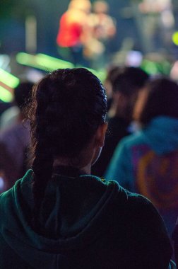 Örgülü saçlı bir insan, canlı sahne ışıkları altında canlı bir konseri dikkatle izleyen kalabalığın arasında duruyor. Festival atmosferi heyecan ve merak yaratır..