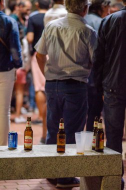 Boş bira şişeleri ve bardaklar canlı bir sosyal toplantı sırasında şehir bankında bırakıldı. Arka planda durup konuşan insanlar vardı..