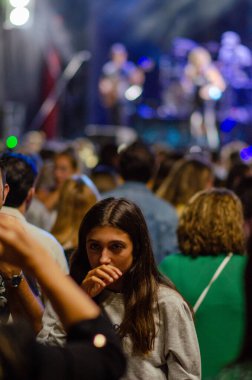 Yansıtıcı genç bir kadın kalabalık bir açık hava konserinin ortasında duruyor, renkli sahne ışıkları ve arka planda canlı performanslar, kentsel gece hayatı atmosferini yakalıyor..