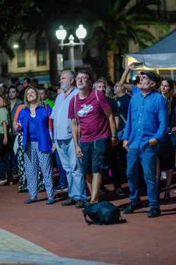Bir grup insan, bir şehir meydanında, gece vakti yaşanan bir olayı dikkatle izliyor, sokak lambalarıyla aydınlatılıyor, meraklarını ve bağlılıklarını dile getiriyorlar..