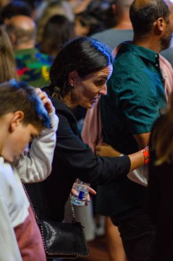 Kalabalık bir etkinlikte farklı insanların etkileşime geçtiği canlı bir kapalı mekan sahnesi. Elinde su tutan, sosyal atmosfer ve bağlantı gösteren siyah ceketli kadına odaklan..