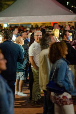 Akşamları açık hava festivalinde sosyalleşen bir grup yetişkin. Parlak ışıklar, günlük kıyafetler ve rahat atmosfer şehir gece hayatı kavramını yakalar..