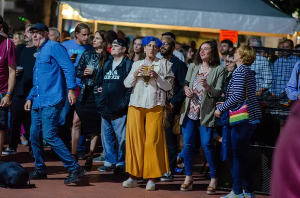 Bir grup yetişkin açık hava festivalinde ayakta durup canlı müzik dinliyor. Şehir eğlencesi, etkinlik atmosferi ve sosyal toplanma sahnesi.