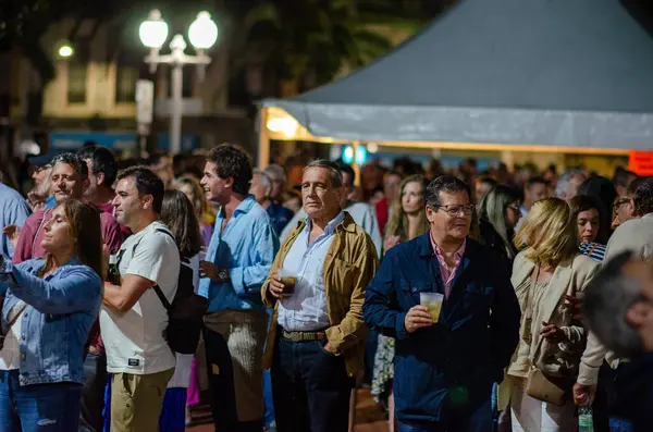 Yetişkinlerden oluşan büyük bir grup, bir açık hava festivalinde bir araya gelerek, içki içiyor ve sohbet ediyor, canlı bir topluluk etkinliğinin atmosferini yakalıyor..