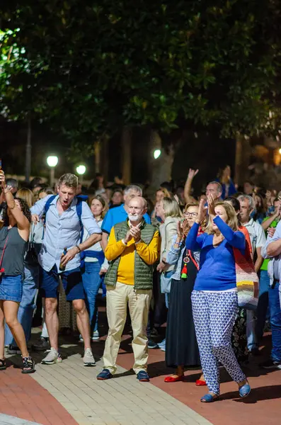 Canlı çeşitlilikteki yetişkinler gece dışarıda toplandı, bir şehir parkında heyecan ve olumlu duygularla canlı eğlence izlediler ve alkışladılar..