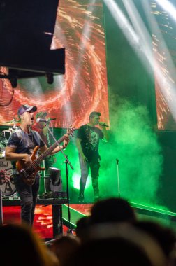 Canlı ışıklar ve yeşil dumanla çevrili bir rock grubunun canlı performansı dinamik müzik atmosferini ve seyirci heyecanını yakalıyor..