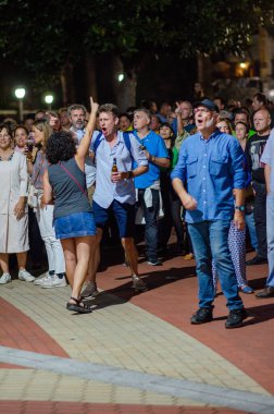 Canlı bir grup insan gece dışarıda toplandı, gülümseyerek, şarkı söyleyerek ve kutlama yaparak bir festival şehir etkinliği sırasında. Şehir gece hayatı, aktif kalabalık, neşeli atmosfer.