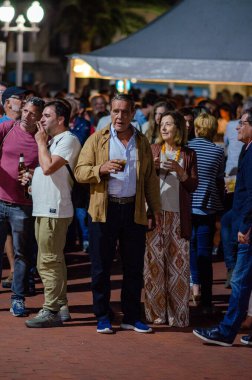 Kalabalık bir grup yetişkin canlı bir açık hava festivalinde ya da sokak partisinde içki içip eğlenerek neşeli bir atmosfer yaratıyor..