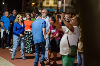 Akşamları açık hava sokak etkinliğinin keyfini çıkaran bir grup yetişkin, gülümseyen, fotoğraf çeken ve rahat bir şehir atmosferinde sohbet eden insanlar..
