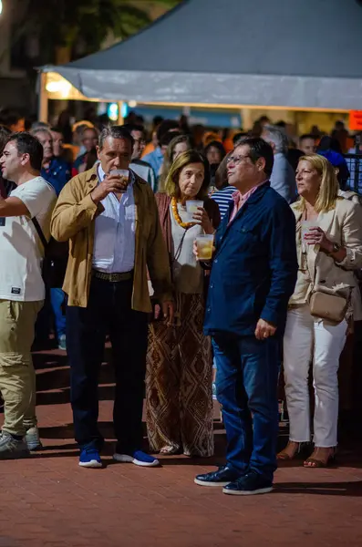 Bir grup insan, açık hava festivalinde çadırın altında içip kaynaşıyorlar. Sıradan sohbetler ve hareketli gece piyasası ışıklarıyla dostane bir sosyal atmosfer..