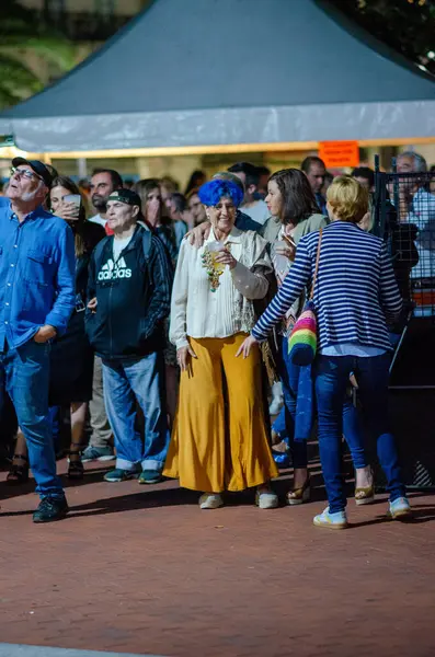 Çeşitli bir grup insan, mavi saçlı ve canlı modaya sahip gülümseyen yaşlı bir kadının yer aldığı açık hava festivalinin keyfini çıkarıyor. Sosyal toplanma, gece hayatı atmosferi.
