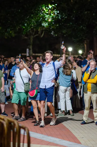 Bir açık hava etkinliğinde kutlama yapan, içki içen ve heyecan gösteren bir grup insan. Canlı gece hayatı, şehir festivali ve sosyal toplanma atmosferini yakalar.
