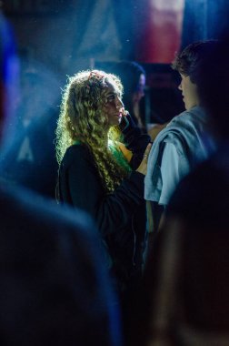 Canlı, renkli bir kulüp atmosferinde akıllı telefondan konuşan kıvırcık saçlı genç bir kadının portresi. İfade dolu gece hayatı duyguları, kentsel gençlik yaşam tarzları..
