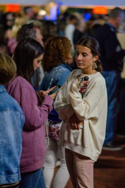 İki genç kadın, canlı bir şehir kalabalığının ortasında, biri telefonunu kontrol ederken diğeri de anahtarlarını tutarken, rahat ve modern bir sosyal atmosferi paylaşıyor..