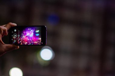 Bir kişi canlı bir açık hava müzik festivalinde akıllı telefonuyla renkli sahne ışıkları ve şehir atmosferiyle bir konser veriyor..