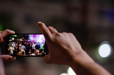 Bir kişi, canlı bir kalabalık ve şehir ışıkları ile çevrili akıllı telefonuyla canlı bir gece konseri kaydediyor. Gece hayatını yakalamak, canlı olay atmosferi.