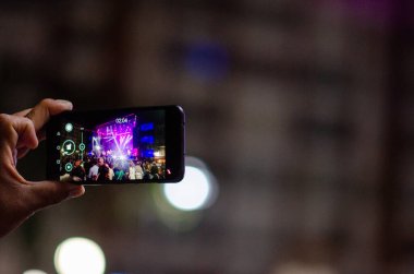 Canlı müzik konserini kaydeden akıllı telefonu tutan bir elin yakın çekimi. Parlak sahne ışıkları kalabalığı aydınlatıyor, heyecan ve gece hayatı atmosferini aktarıyor.