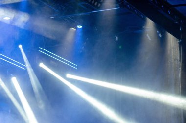Parlak beyaz ışıklar sahne içinde konser dumanının arasından çarpıcı ışınlar oluşturur. Modern bir olay alanında enerji ve heyecan uyandırır..