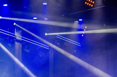 Sahne ışıklandırması ve mavi spot ışıkları bir etkinlik salonunda dumanı aydınlatarak, canlı bir parti veya konser atmosferi yaratıyor..